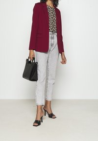Blazer bordeaux sopra una blusa floreale, abbinato a jeans chiari a vita alta e sandali neri con tacco. Una borsa nera con texture completa il look.