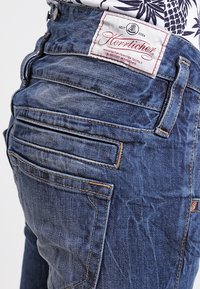 Detailní záběr na zadní kapsu modrých džínsů s hnědým šitím a bílou textilní etiketou s nápisem "Herrlicher", přišitou na pase.