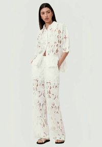 Witte kanten twee-delige outfit bestaande uit een kort-lang mouwige button-up shirt en wijde broek, met ingewikkelde bloemvormige uitsnijdingen.
