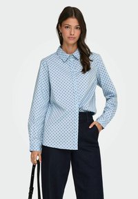 Chemise à pois bleu clair en coton, avec un col et une fermeture à boutons, portée avec un pantalon en denim foncé, assortie à un sac à bandoulière noir.
