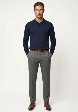 Mand står med hænderne i lommerne iført marineblå langærmet polo, grå bukser med brunt bælte og brune lædersko på en ensfarvet baggrund.