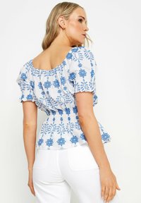 Hvid bluse med blå blomsterbroderi, korte puffærmer og smocket talje. Bagsiden har en bred halsudskæring og flæset kant.