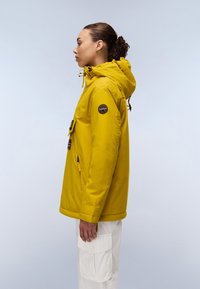 Gelbe wasserdichte Jacke mit Kapuze, mit einer Vordertasche, Reißverschlussakzenten und einem Logopatch. Getragen mit weißen Cargo-Hosen.