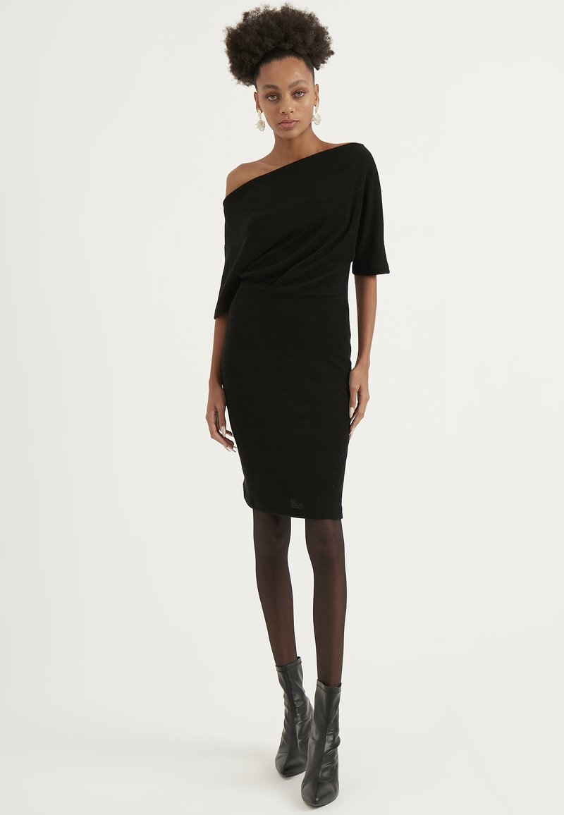 Abito nero a coste con design off-shoulder, maniche corte e silhouette aderente. Abbinato a stivali neri al ginocchio.