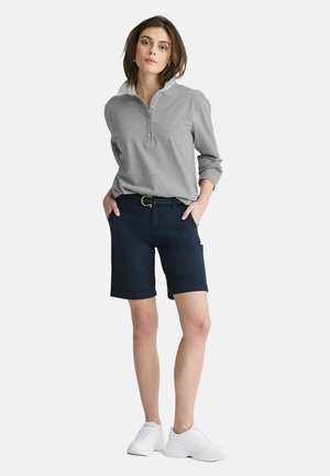 Grå langærmet top med krave og knapper, parret med marineblå shorts. Hvide sneakers fuldender outfittet og fremhæver en afslappet stil.