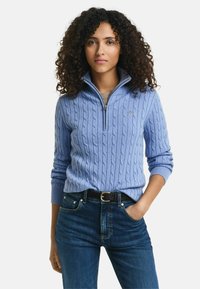 Helles blaues Kabelstrickpullover mit einem Halb-Reißverschlusskragen, gerippten Bündchen und einem kleinen Logo auf der Brust, kombiniert mit dunkelblauen Jeans.