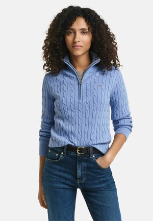 Light blue cable-knit sweater with a half-zip collar, ribbed cuffs, and a small logo on the chest, paired with dark blue jeans.
