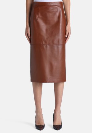 Jupe crayon en cuir marron longueur genou portée avec des chaussures à talons hauts et bouts pointus sur une personne debout devant un fond uni.