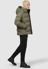 Chaqueta acolchada verde olivo con capucha, frente con cremallera y bolsillos laterales, combinada con pantalones negros y botines negros. Textura suave.