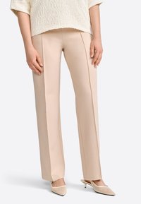 Pantalon beige ajusté avec une texture lisse, une coupe droite et des coutures avant discrètes, associé à des mules en maille à talons bas.