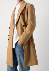 Cappotto in lana color cammello con design doppiopetto, bottoni neri e vestibilità leggermente oversize. Abbinato a una camicia bianca e jeans blu.