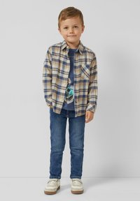 Kariertes Flanellhemd in Beige und Marineblau über einem dunklen T-Shirt mit Grafik, kombiniert mit blauen Jeans und weißen Sneakern mit beigen Akzenten.