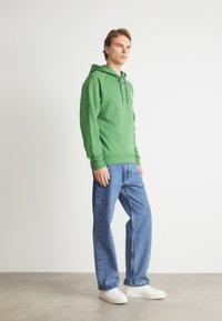 Πράσινη μπλούζα με κουκούλα και κορδόνια, σε συνδυασμό με ανοιχτό μπλε τζιν και λευκά sneakers. Η μπλούζα διαθέτει άνετη εφαρμογή και μαλακή υφή.