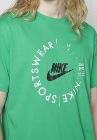 Vihreä puuvillapaita, jossa on musta Nike-logo, pyöreä valkoinen teksti ja graafinen kuvasto, johon sisältyy koordinaatit ja lämpötilasymboli.