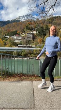 Camisola atlética azul-claro de manga comprida, leggings pretos e sapatilhas brancas. Passadiço pavimentado ao lado de um rio verde com folhagem de outono ao fundo.