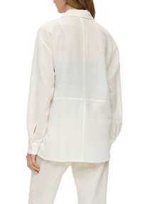 Vit linneskjorta med krage, långa ärmar och sidoklyftor. Sömmar synliga på baksidan, lätt och något genomskinlig textur.