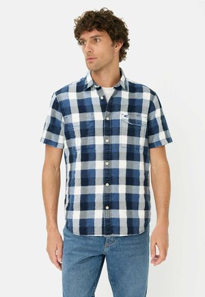 Mand iført kortærmet skjorte med marineblåt, blåt og hvidt ternet mønster over hvid T-shirt og denimjeans foran en ensfarvet hvid baggrund.