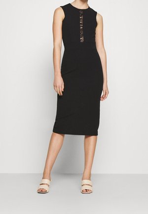 Vrouw draagt een mouwloze zwarte midi-jurk met verticale kanten details op de borst, gecombineerd met beige open-toe instapsandalen.