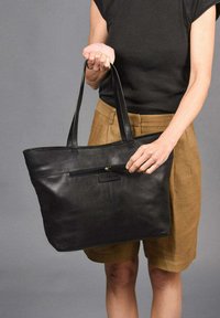 Bolso tote de cuero negro con asas largas, que cuenta con un bolsillo frontal con cremallera y una textura suave. Diseño simple y rectangular con un detalle de marca mínimo.