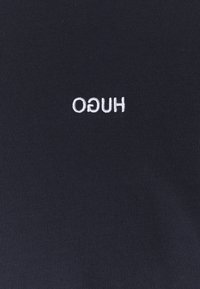 Schwarzes Stoff-T-Shirt mit weißem gesticktem "HUGO"-Logo, das eine glatte Textur und ein engmaschiges Design zeigt.