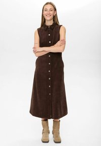 Rjavo kordurojno brezrokavno obleko z gumbnim zapenjanjem spredaj, ovratnikom, stranskimi žepi in dolžino do meč, v kombinaciji s bež gležnjarji.
