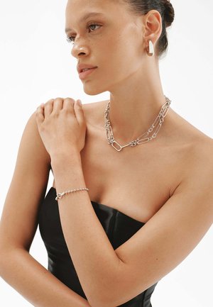 Collar de cadena de plata con eslabones, pulsera a juego, llevada por un modelo en la parte superior del cuerpo, con texturas suaves y un diseño minimalista.