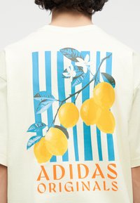 Άτομο που φοράει κρεμ πουκάμισο με μπλε κάθετες ρίγες, απεικόνιση κίτρινων λεμονιών και λευκών λουλουδιών, πορτοκαλί κείμενο «Adidas Originals» στην πλάτη.