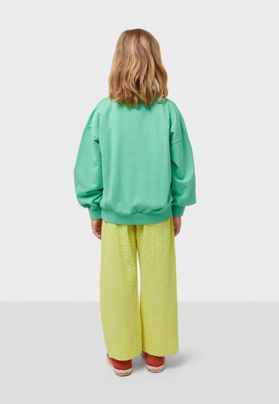Felpa verde con maniche gonfie, abbinata a pantaloni a gamba larga gialli con texture. Modella indossa scarpe arancioni, vista da dietro.