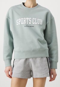 Felpa verde chiaro con la scritta "SPORTS CLUB" e "BJÖRN BORG" stampata in bianco. Tessuto morbido, vestibilità ampia e polsini a coste; abbinata a pantaloni corti grigi.