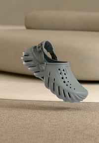 Γκρι αεριζόμενο τσόγκ με σόλα κυματοειδούς σχεδίου και ρυθμιζόμενο λουράκι στη φτέρνα, αιωρούμενο πάνω από ένα μπεζ χαλί σε ένα ουδέτερο δωμάτιο.