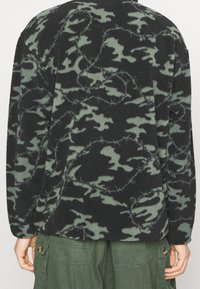 Schwarze und grüne Camouflage-Fleecejacke mit weicher Textur, langen Ärmeln und lockerem Sitz. Auffällige Nähte entlang der Nähte.