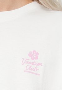 Hvid bomuld t-shirt med en lyserød "Vacation Club"-logo og et hibiscusblomst design over logoet. Glat tekstur, standard rund halsudskæring.