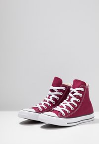 Burgunderfarbene High-Top-Sneaker aus Canvas mit weißen Schnürsenkeln, Gummispitze und schwarzem Streifen entlang der Sohle. Glatte Textur, klassisches Design.