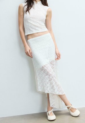 Nainen pukeutunut hihattomaan valkoiseen pitsiseen crop-topiin, epäsymmetriseen valkoiseen pitsihameeseen ja valkoisiin balleriinakengiin, seisomassa valkoista seinää vasten.