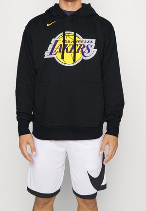 Homme portant un sweat à capuche noir des Los Angeles Lakers avec un logo jaune et violet, et un short de basketball blanc Nike avec un swoosh noir.