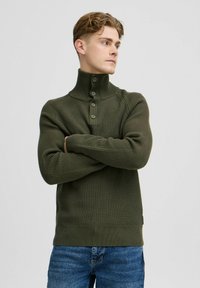 Olivgrüner gerippter Strickpullover mit hohem Kragen, ausgestattet mit drei Knöpfen und einer figurschmeichelnden Passform. Kombiniert mit blauen Jeans.