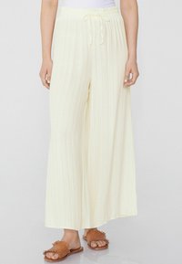 Pantalones anchos de color amarillo claro con rayas verticales, cintura elástica y cordón ajustable. Combinados con sandalias de tiro marrón con flecos.