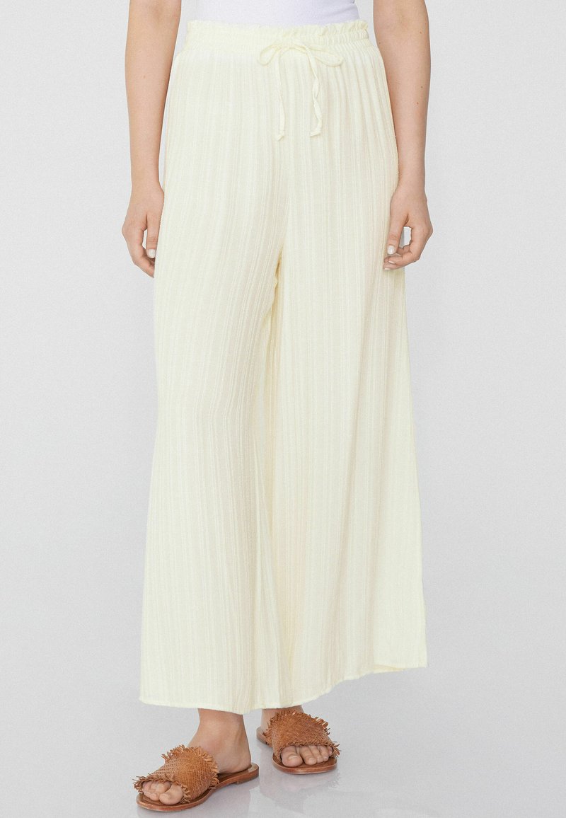 Pantalones anchos de color amarillo claro con rayas verticales, cintura elástica y cordón ajustable. Combinados con sandalias de tiro marrón con flecos.