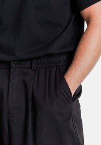 Zwarte elastische broek met een losse pasvorm, voorzien van zijzakken en een gladde, gestructureerde stof. Korte zwarte top die gedeeltelijk zichtbaar is.