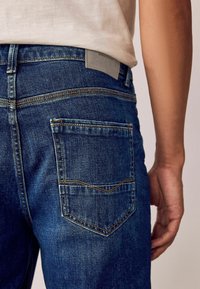 Jeanși din denim de culoare albastră închisă, cu o textură netedă. Au cusături aurii și o etichetă pe talie intitulată "DENIM SUPPLY". Designul buzunarului din spate este înclinat.