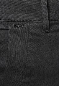 Pantalones de mezclilla negros con una superficie texturizada, que cuentan con una cinturilla cosida y un pequeño logotipo RAW grabado en el bolsillo delantero.