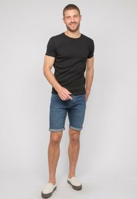 T-shirt noir à manches courtes, short en denim bleu avec des revers, et chaussures slip-on beige. Design sobre, coupe décontractée et matériaux légers.