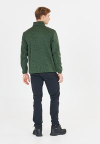 Grøn, tekstureret pullover med høj krave, parret med sorte bukser og støvler. Har et enkelt design uden synlige logoer.
