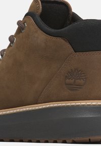 Brown suede shoe with a smooth texture, featuring gray accents, metal eyelets, and a logo embossed on the side. Rubber sole with white stitching.
