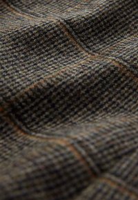 Nærbillede af struktureret uldstof med hundetandsmønster i mørkegrøn og brun, med tynde orange rudemønstrede linjer.