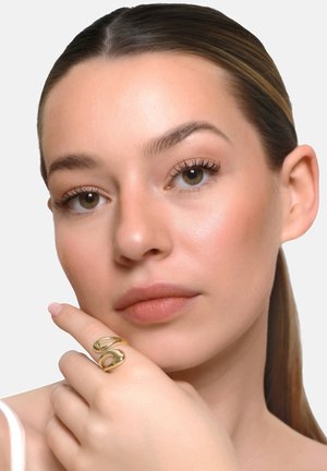 Donna con pelle liscia, capelli scuri raccolti all'indietro, appoggia il mento sulla mano indossando un anello moderno e spesso in oro sull'indice.