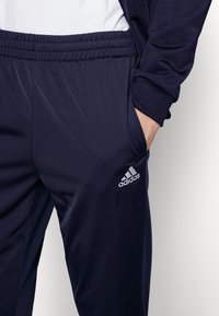 Pantalones de chándal azul marino con cintura elástica, textura suave, un bolsillo lateral y un logo de Adidas en plata reflectante.