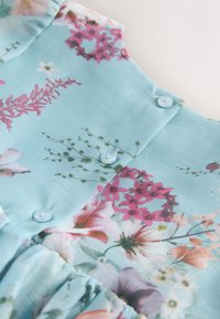 Tissu floral bleu clair avec des fleurs roses, blanches et violettes, présentant de petits boutons et un détail de couture froncée.