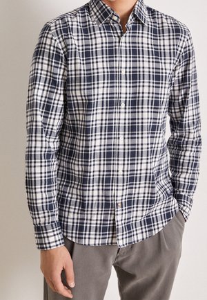 Mies, joka käyttää laivastonsinistä ja valkoista ruudullista pitkähihnaista nappipaitaa ja harmaita housuja, seisoo käsivarsi taskussa.