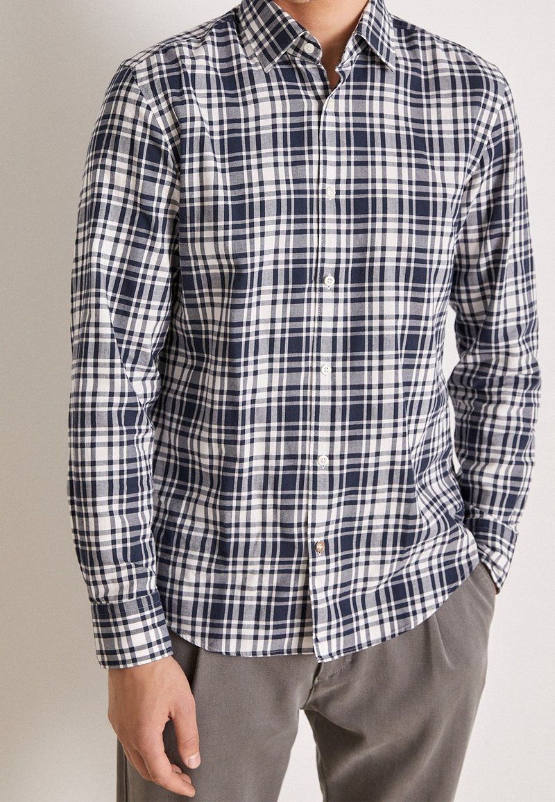 Homme portant une chemise à carreaux bleu marine et blanc à boutons avec manches longues et un pantalon gris, debout avec une main dans la poche.
